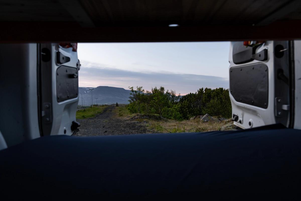 Blick vom Bett durch die geöffneten Hecktüren auf einen Schotterweg und die Berge