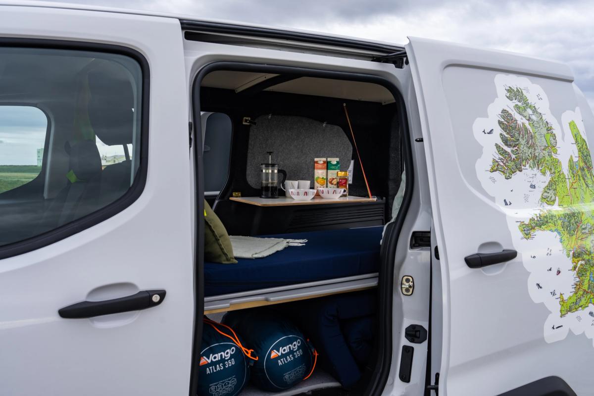 Puerta lateral corredera abierta junto a la cama, mesa con productos para el desayuno y sacos de dormir guardados debajo