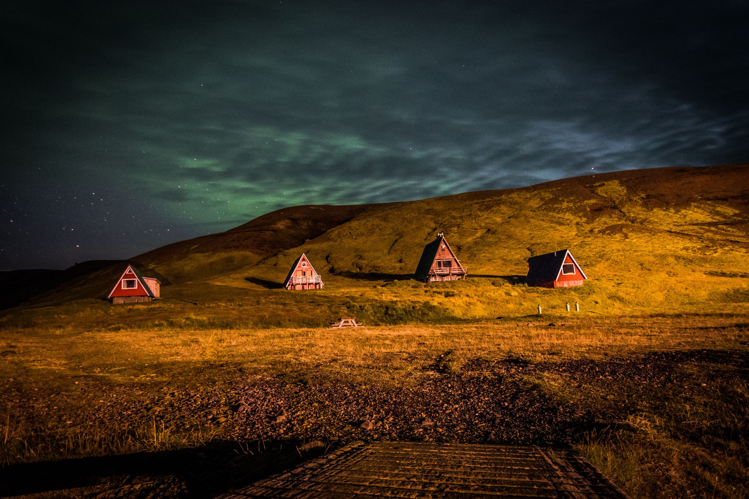 Cuatro cabañas con estructura en A y techos rojos se sitúan sobre colinas doradas bajo un cielo nocturno con aurora boreal verde.