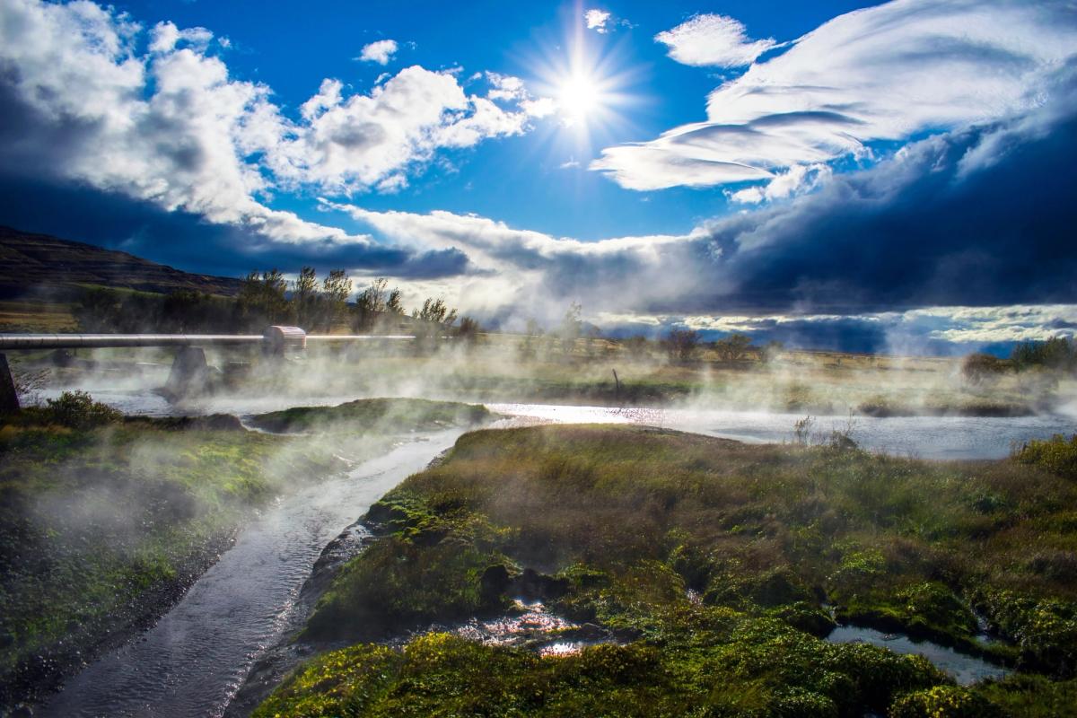 A sunlit, misty river landscape with a pipe bridge crossing in the background under a partly cloudy sky.