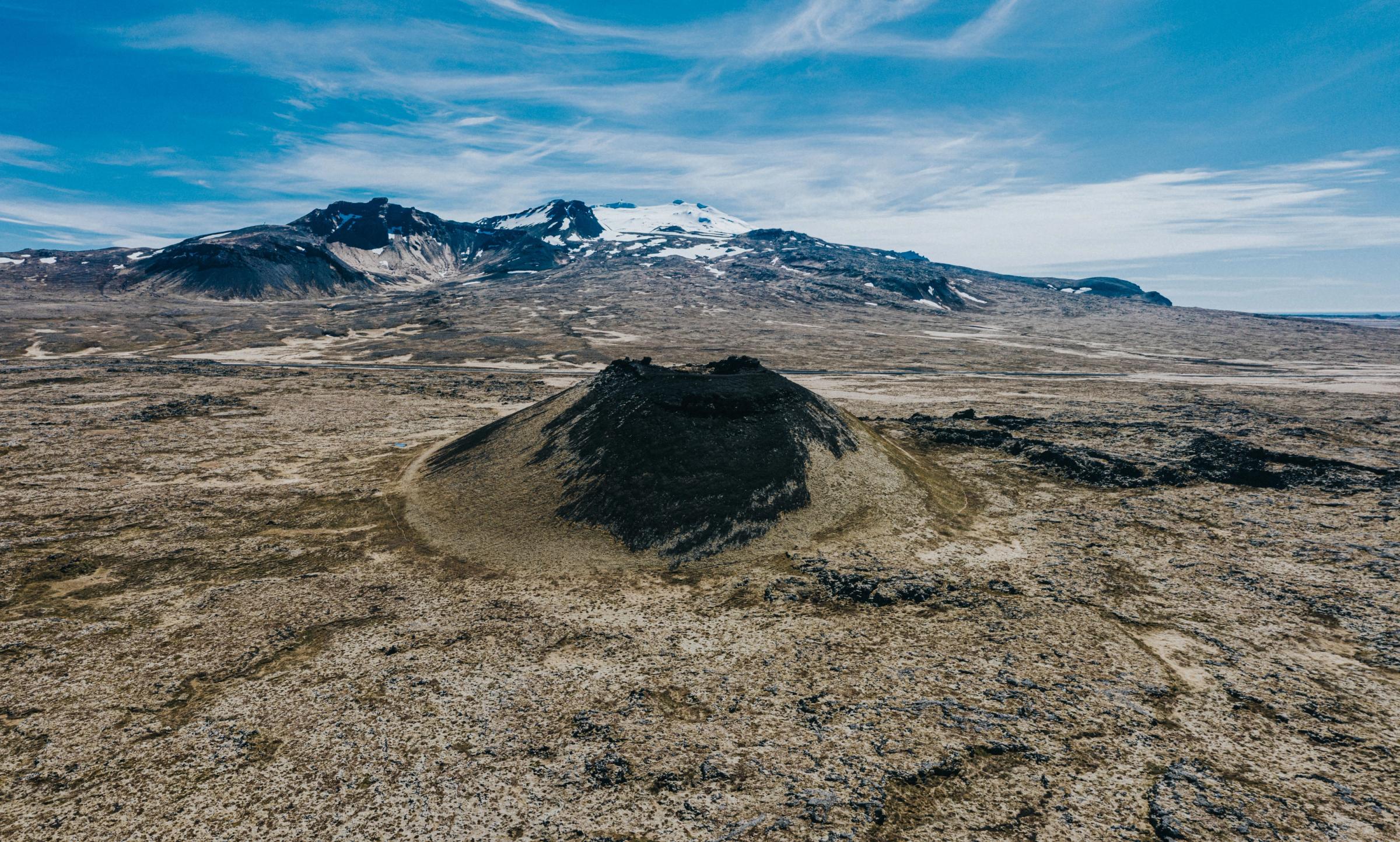 Dark volcanic cone in a rocky lava field with snow-capped mountains under a blue sky.