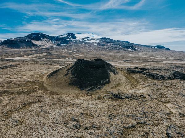 A dark volcanic cone in a barren lava field, with snow-capped mountains under a blue sky.