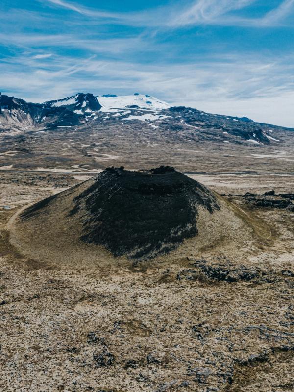A dark volcanic cone in a barren lava field, with snow-capped mountains under a blue sky.