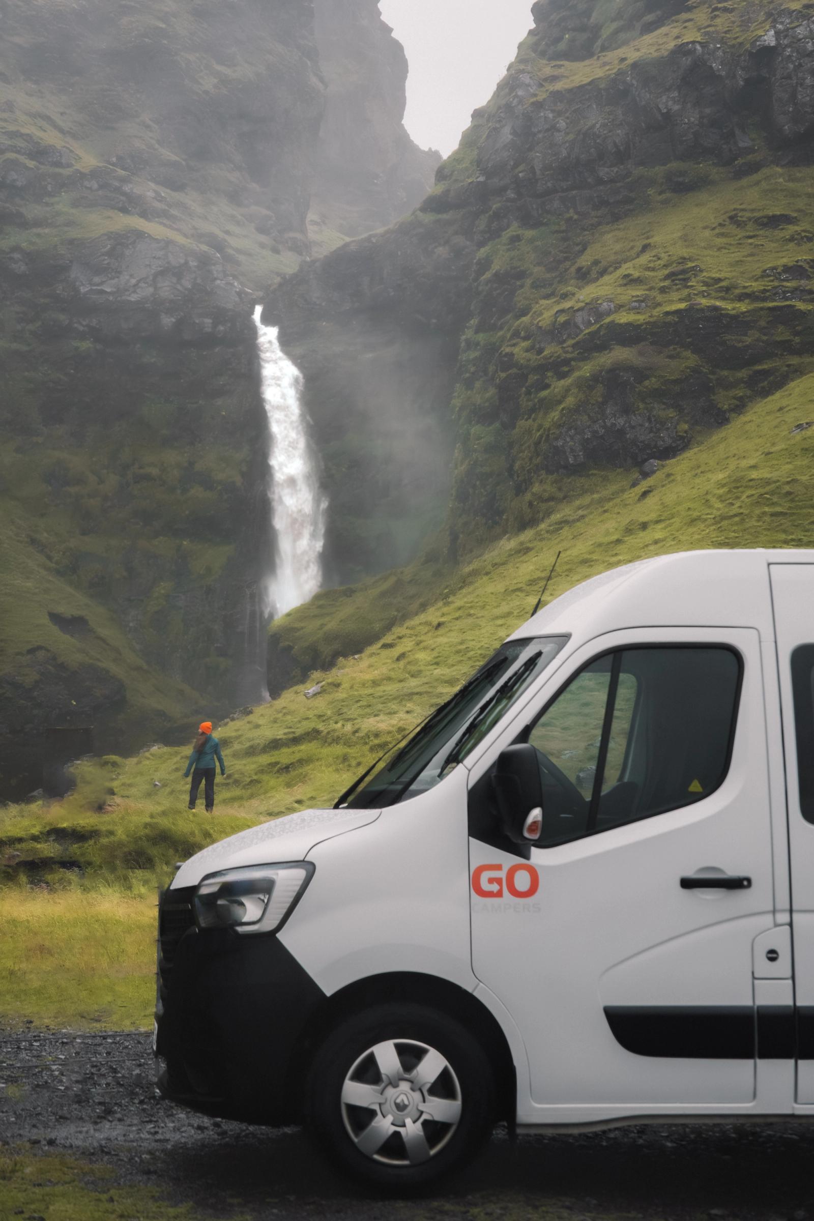 A white camper van parked on the right, with a person viewing a large waterfall in a lush, misty valley.
