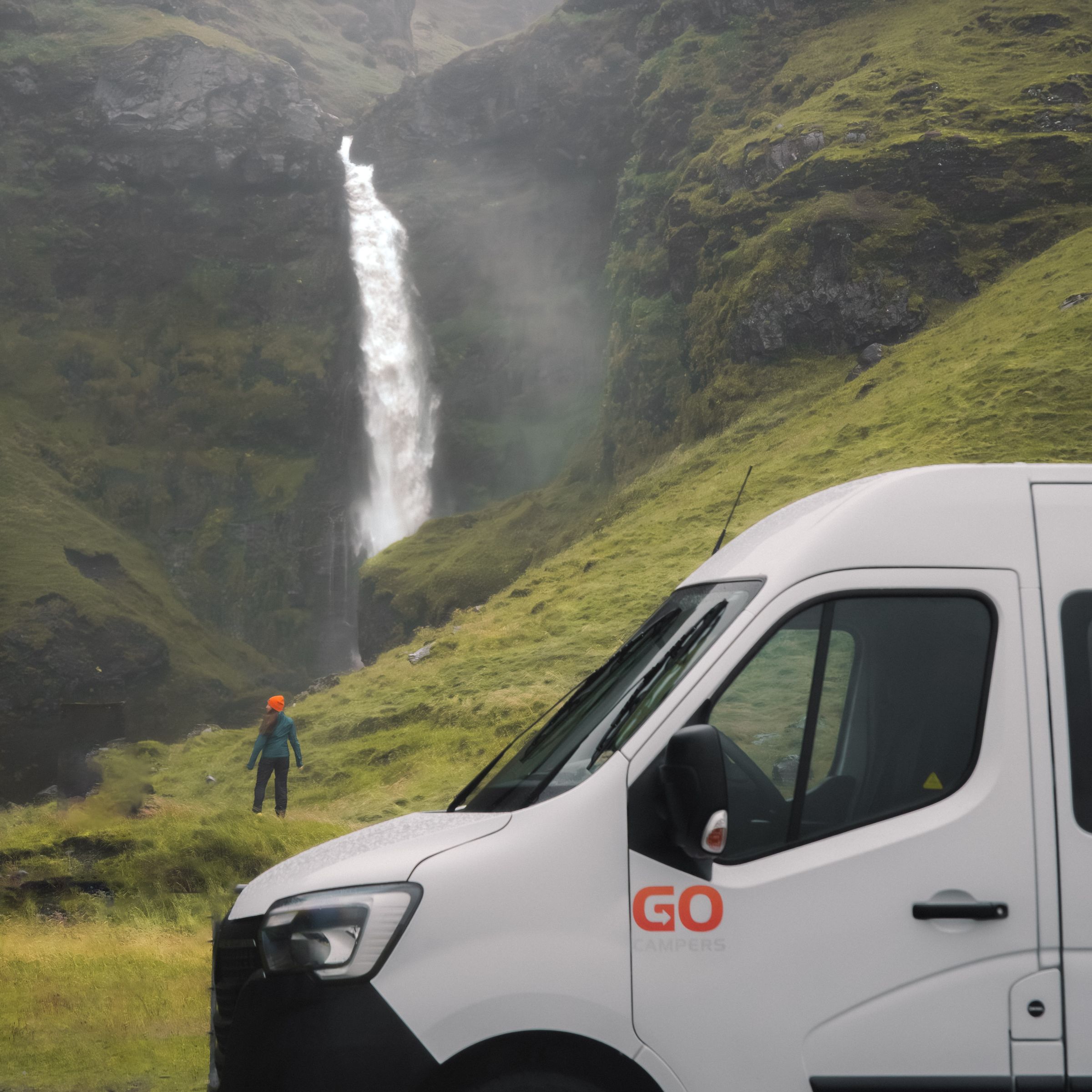 A white camper van parked on the right, with a person viewing a large waterfall in a lush, misty valley.