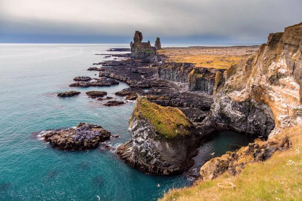 Rugged volcanic coastline with dark rock formations, turquoise water, steep cliffs, and distant sea stacks under a cloudy sky.