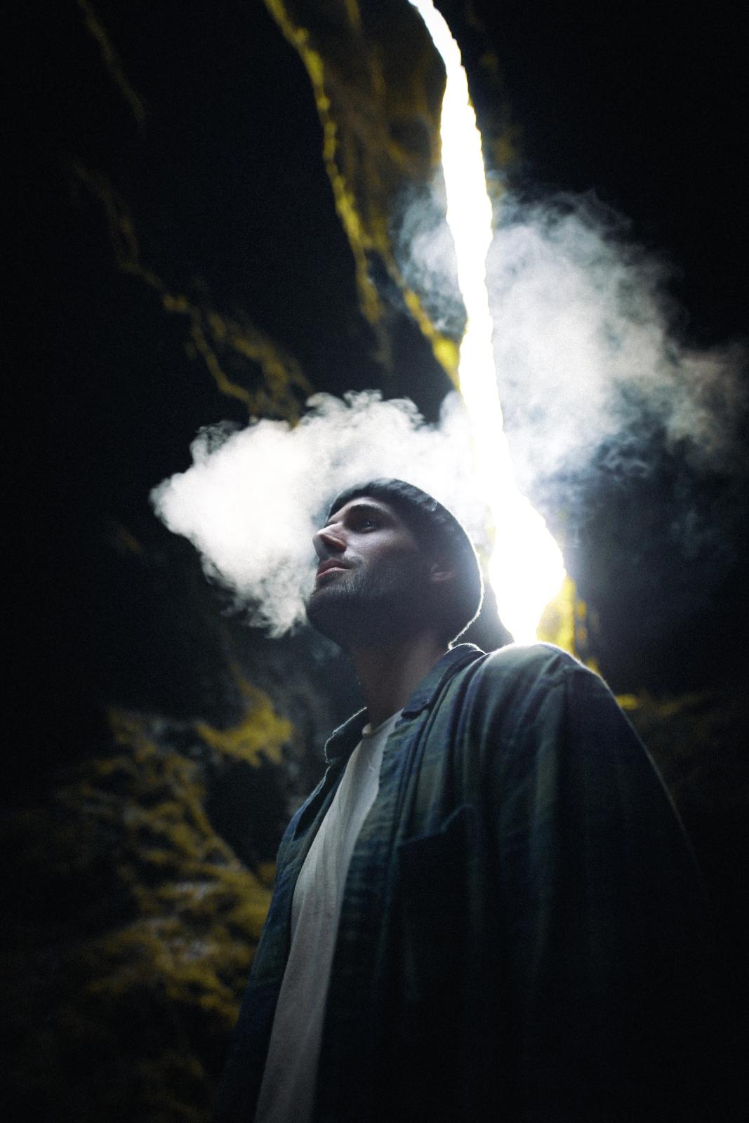 Un hombre con un gorro exhala humo hacia arriba, iluminado por un brillante rayo de luz en una cueva oscura.