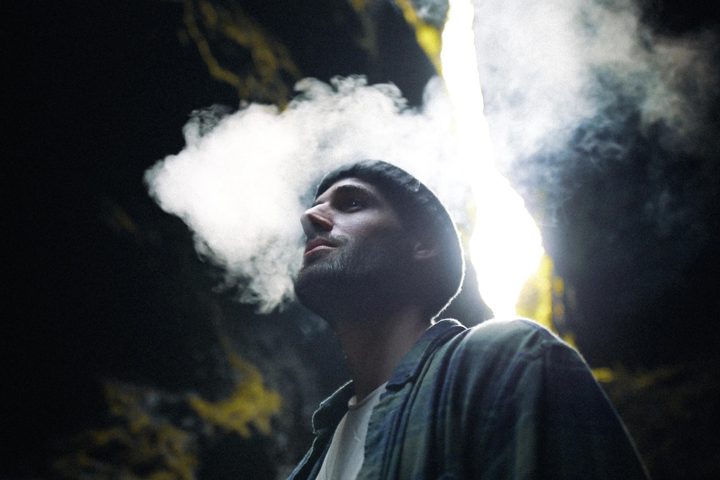 Un hombre con un gorro exhala humo hacia arriba, iluminado por un brillante rayo de luz en una cueva oscura.