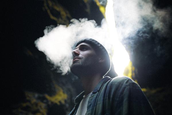 A man in a beanie exhales smoke upwards, illuminated by a bright crack of light in a dark cave.