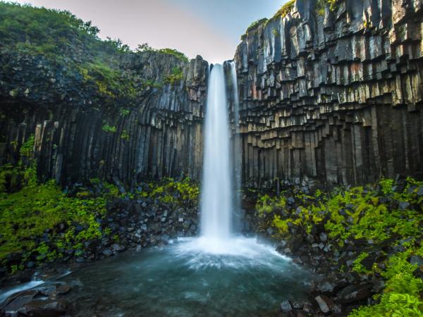 A waterfall cascades over dark hexagonal basalt columns surrounded by green foliage.