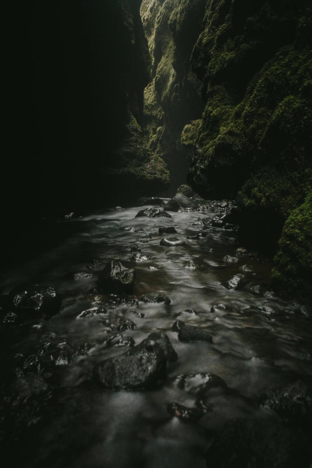 Arroyo fluyendo sobre rocas en un cañón oscuro y musgoso con luz distante.