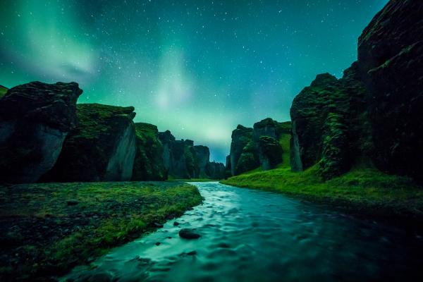 A river flows through a mossy canyon under a starry sky illuminated by green Northern Lights.