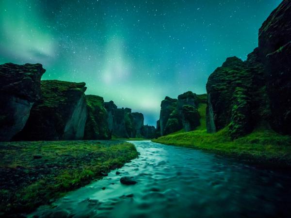 A river flows through a mossy canyon under the green Northern Lights and a starry sky.