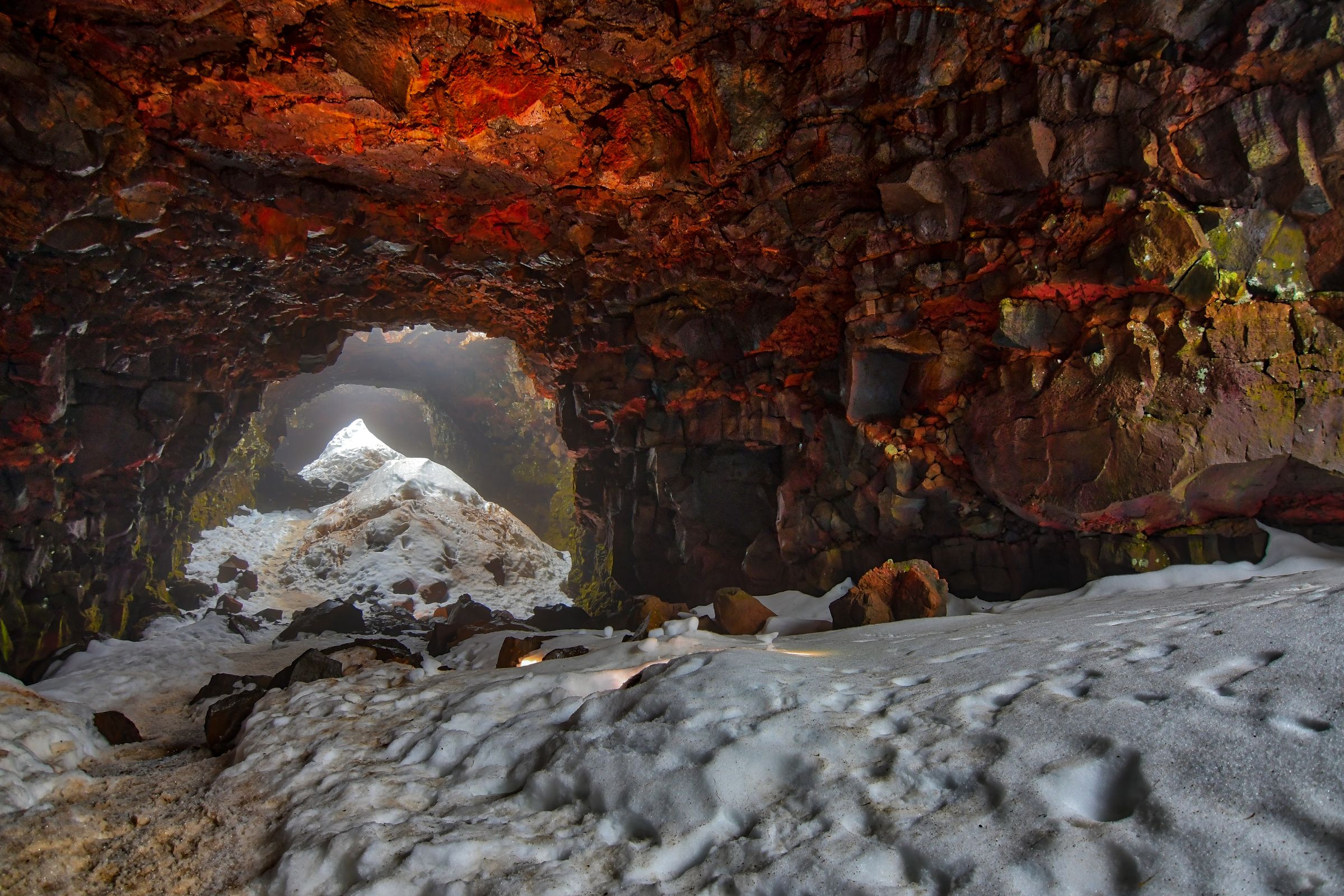 A volcanic cave with reddish rock walls and a snowy floor, opening to a bright exit.