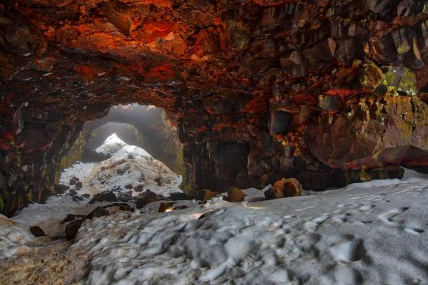 A volcanic cave with reddish rock walls and a snowy floor, opening to a bright exit.
