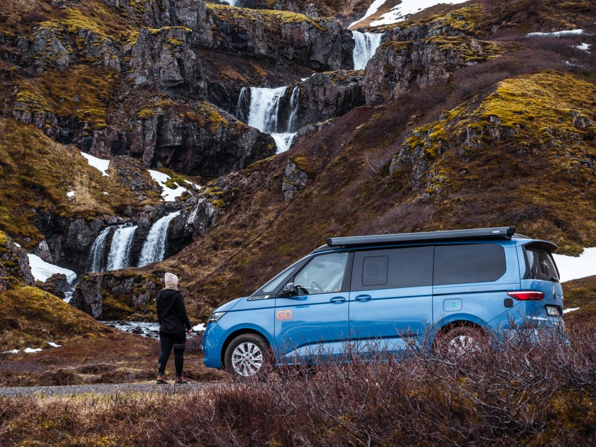 Vue sur la cascade depuis un camping-car 4x4 Un van aménagé 4x4 pour 2 personnes garé au pied de cascades sur des pentes rocheuses, location de van en Islande