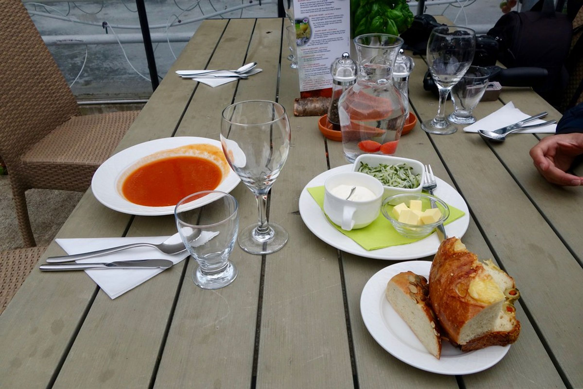 Sopa de tomate, pan, mantequilla, crema y hojas verdes dispuestos sobre una mesa de madera.