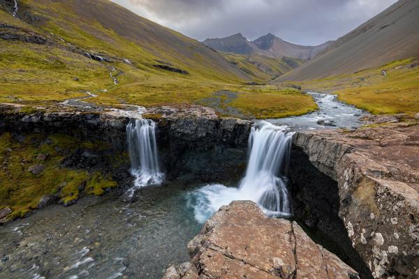 Two waterfalls dropping over a rocky ledge in a scenic valley with a winding river and mountains.