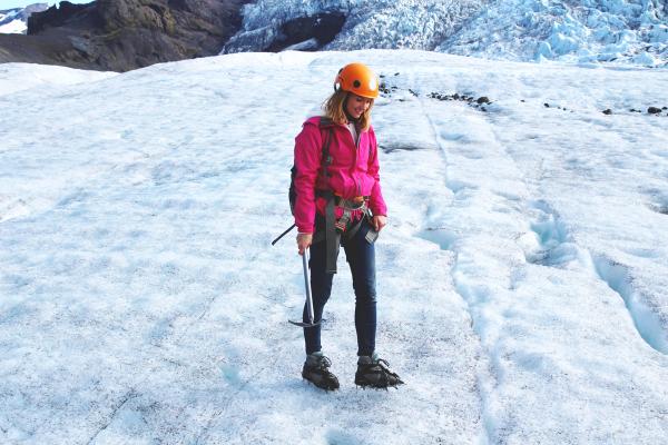 a woman in a pink jacket and orange helmet is walking on a glacier .