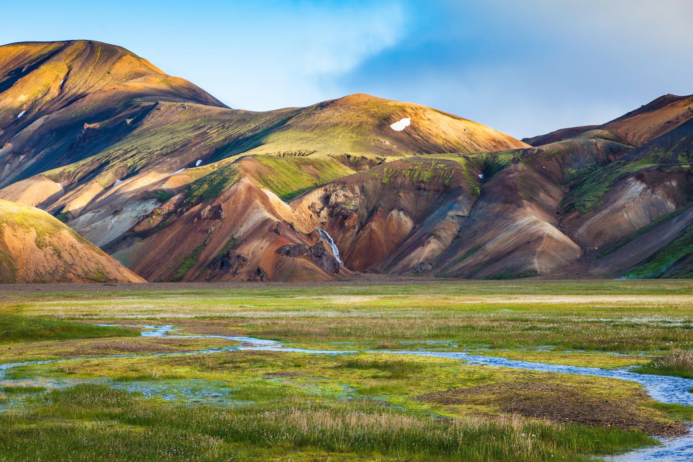 Colorful mountains with a waterfall, overlooking a green valley with a winding stream.