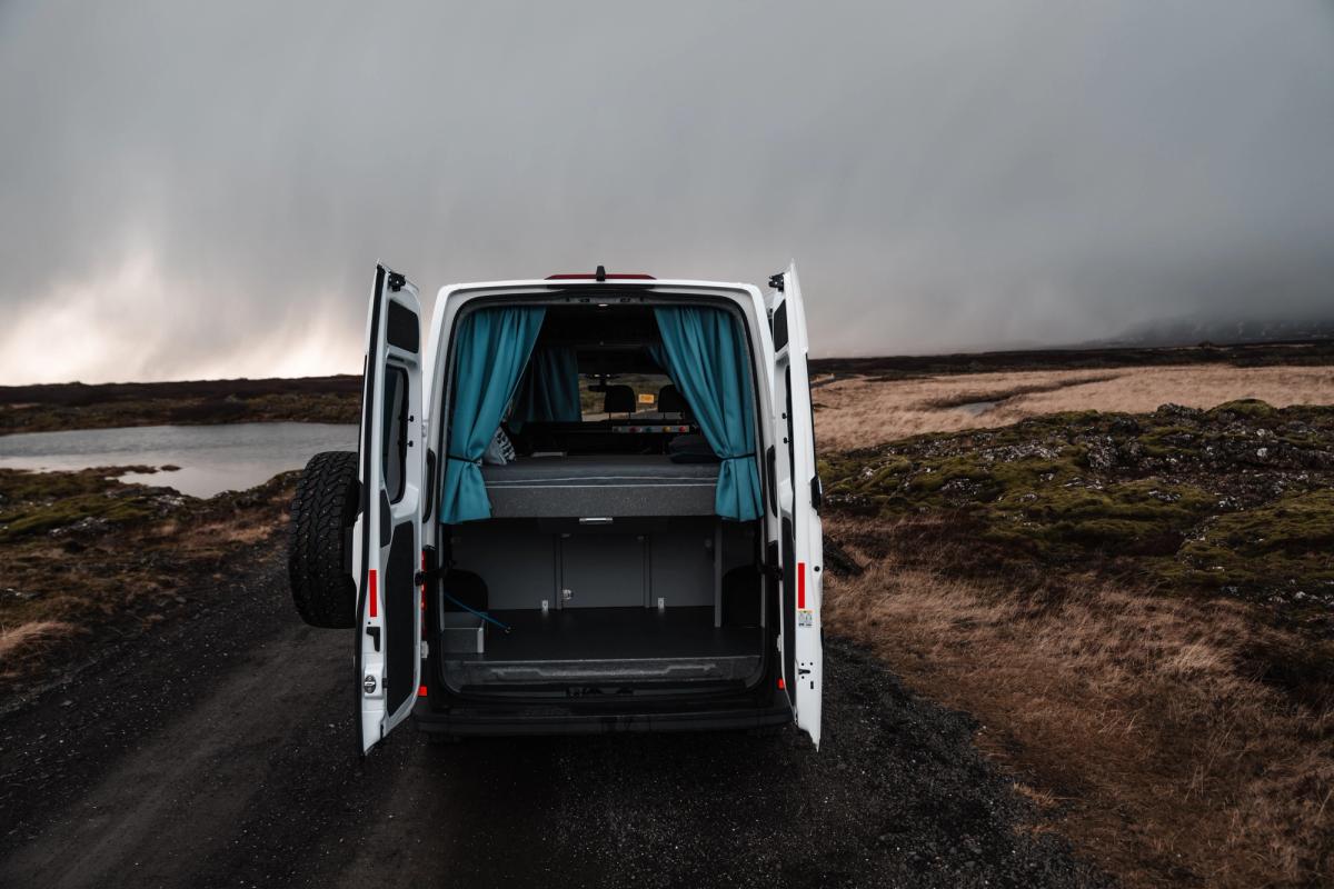 Las puertas traseras abiertas dejan ver la cama y el espacio de almacenamiento inferior de una furgoneta cámper en un camino de grava en Islandia
