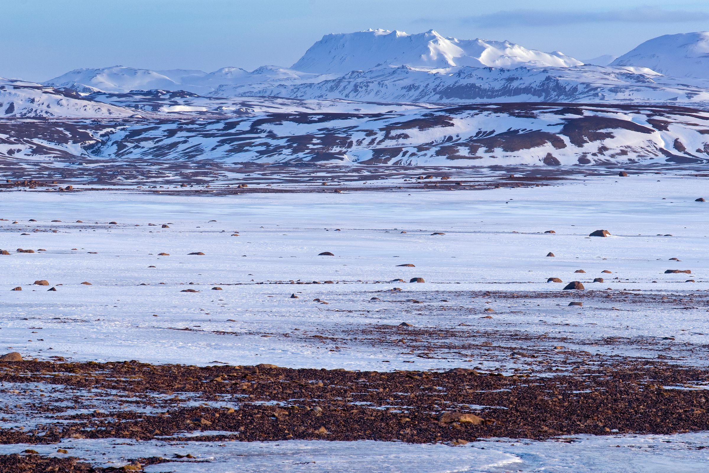 A wide, snow-covered landscape with scattered dark ground and distant snowy mountains.