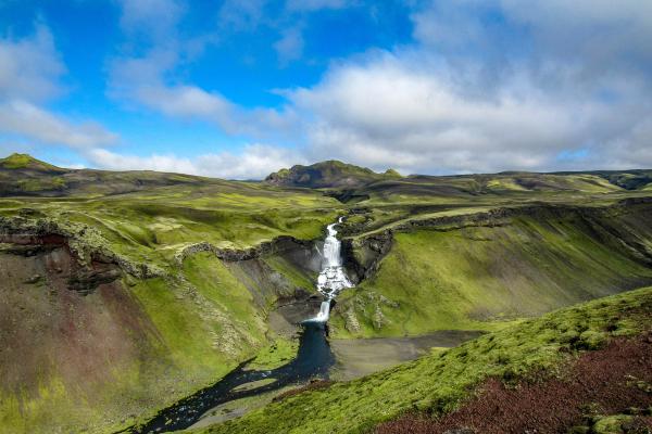 A multi-tiered waterfall cascades into a dark river winding through vibrant green, mossy valleys under a blue, cloudy sky.