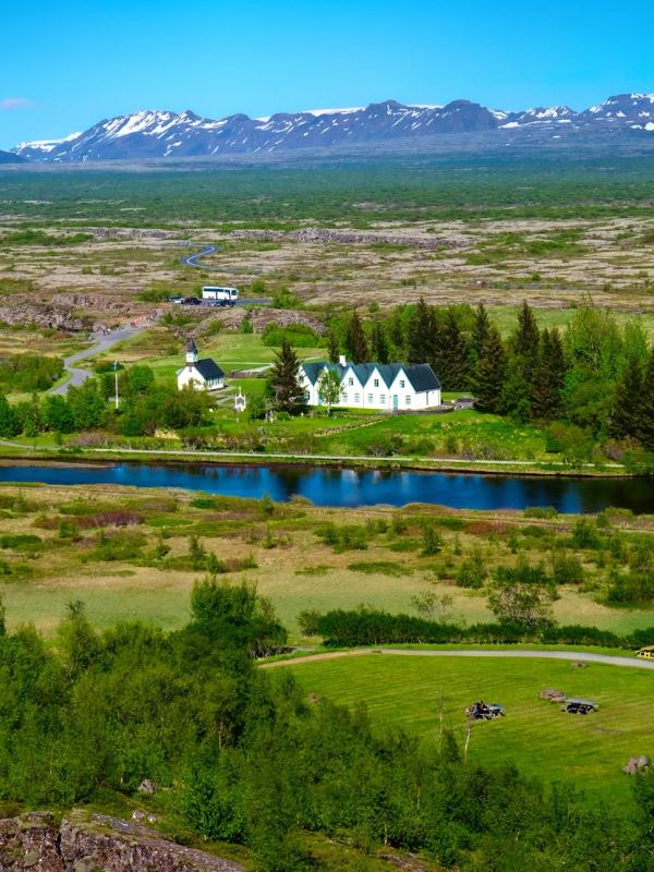Scenic landscape with a winding blue river, white buildings, green fields, and snow-capped mountains.