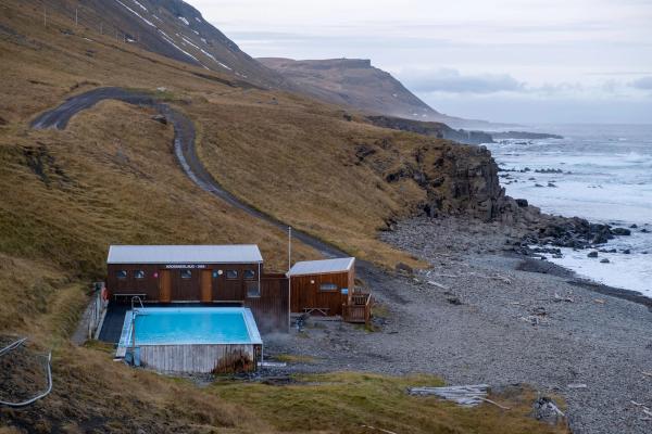 Outdoor geothermal pool and rustic buildings on a rugged, treeless hillside overlooking a rocky ocean coast.