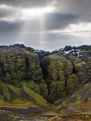 Mossy mountains with a deep crevice under sun-streaked clouds.