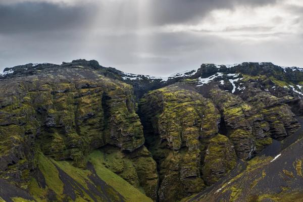 Canyon walls covered in moss under a cloudy sky with a ray of sunlight breaking through.