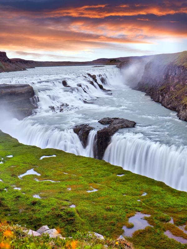 Wide waterfall cascading through a green canyon under a dramatic sunset sky.