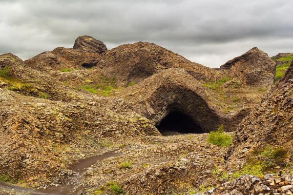 A path leads to a dark cave entrance within a rugged, textured rock landscape under a cloudy sky.