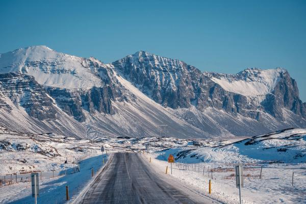 A winter road stretches toward large snow-capped mountains under a clear blue sky.