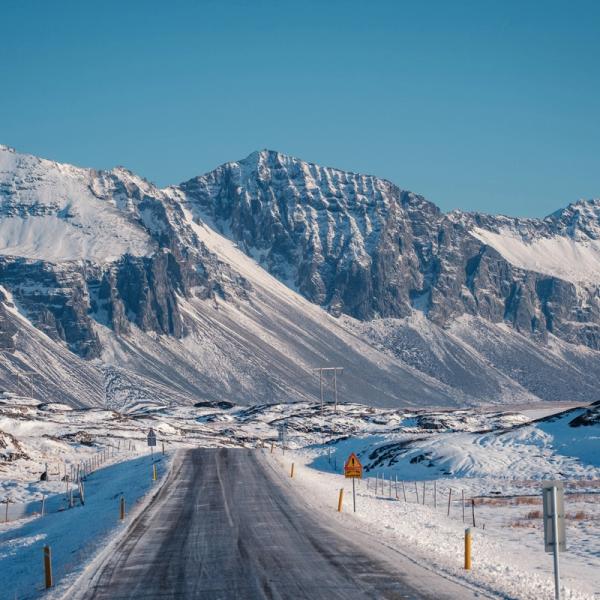 A winter road stretches towards snow-capped mountains under a clear blue sky.
