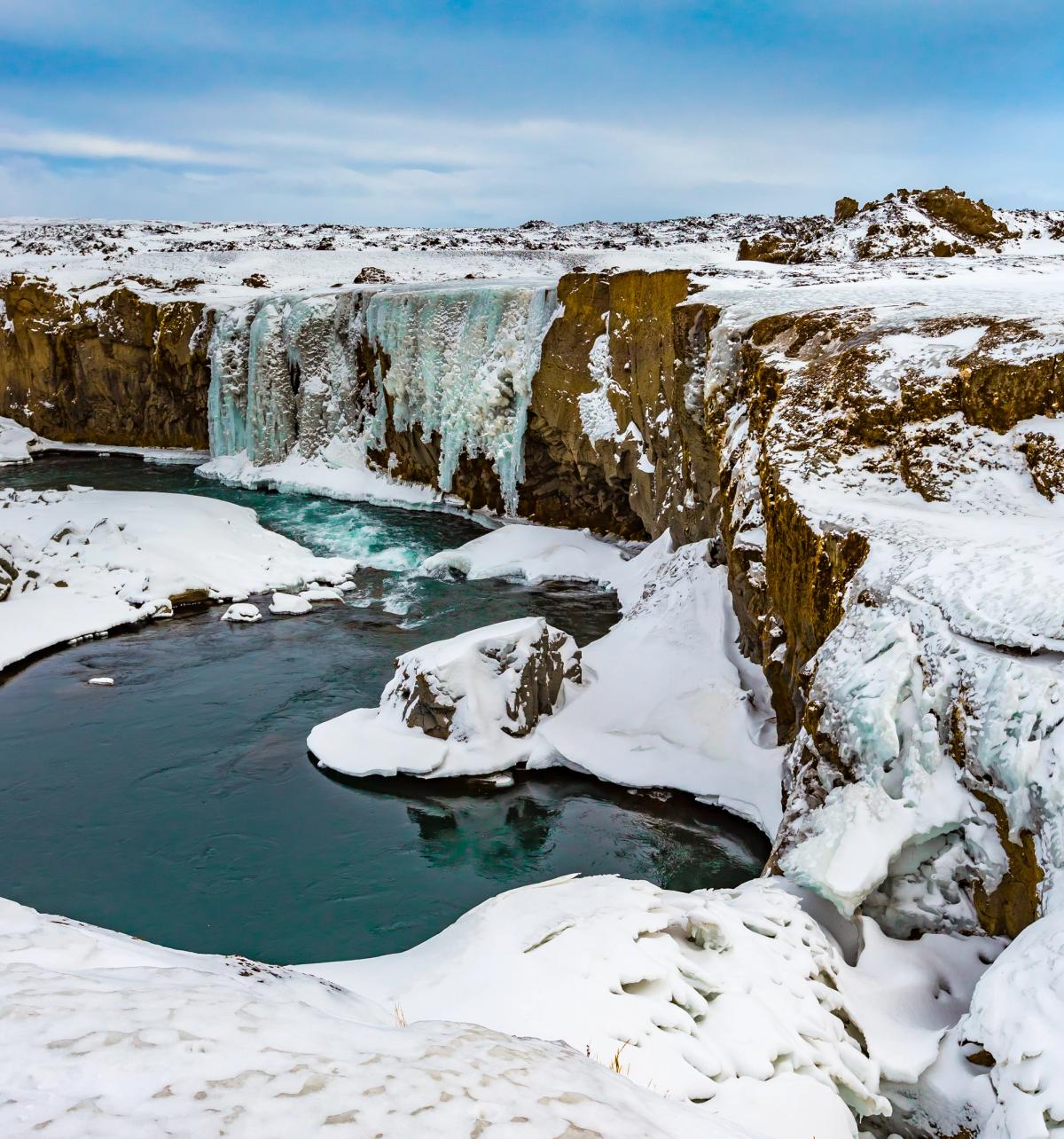 Waterfall with large icicles and a blue river in a snowy canyon.
