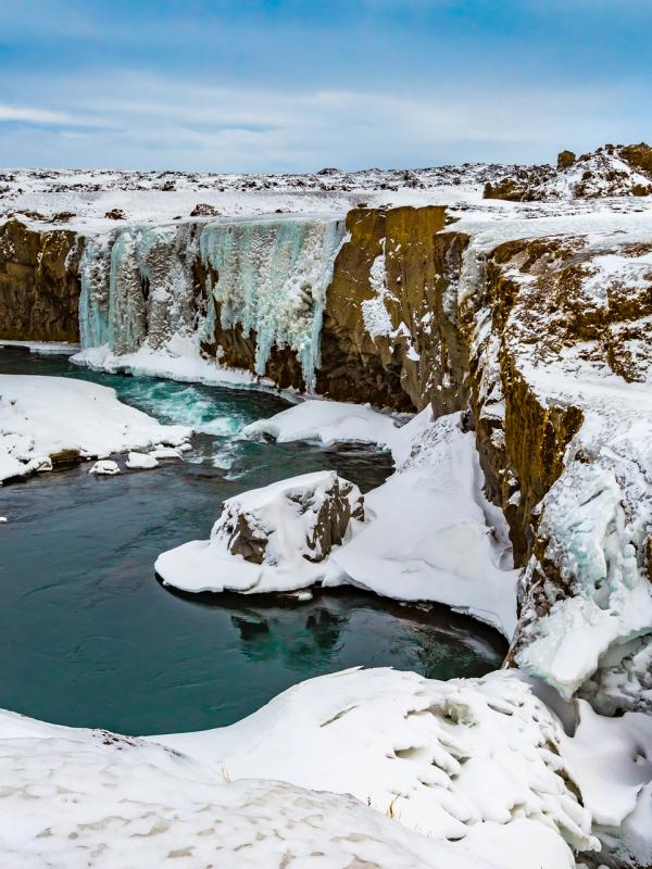 Waterfall with large icicles and a blue river in a snowy canyon.