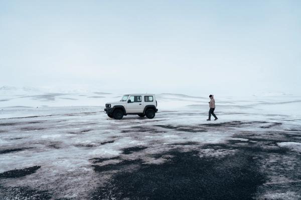 A person walks on dark, patchy snow and ice next to a white SUV in a vast, snow-covered landscape.