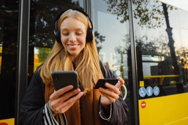 a woman wearing headphones is using a cell phone and a credit card while waiting for a bus .