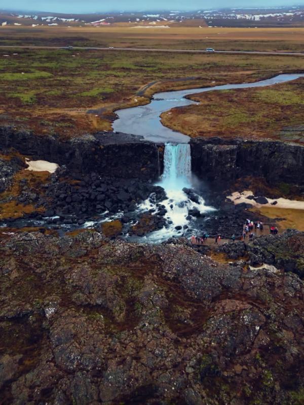 A waterfall cascades between dark rocky cliffs into a river in a vast, rugged landscape, with a few people visible near the falls.