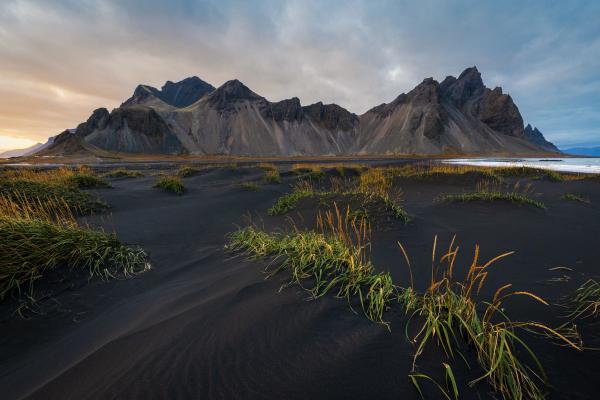 Black sand dunes with sparse grass, jagged mountains, and ocean under a dramatic sky.