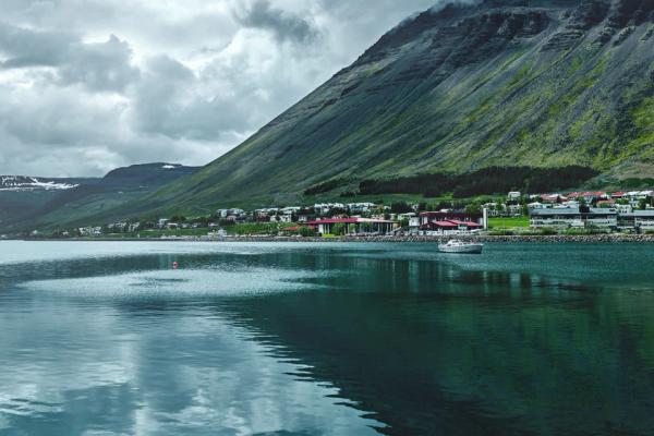 A village sits on a calm bay with a small boat, backed by a large green mountain range under a cloudy sky.