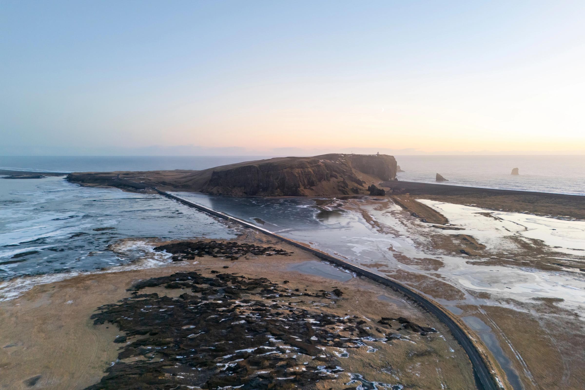 Causeway crossing a partially frozen bay towards a cliff with a lighthouse and sea stacks at sunrise.