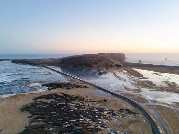 Aerial view of a road crossing icy coastal waters towards a cliff with a lighthouse and sea stacks at sunrise.