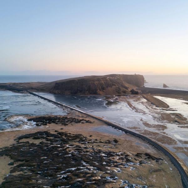 Causeway crossing a partially frozen bay towards a cliff with a lighthouse and sea stacks at sunrise.