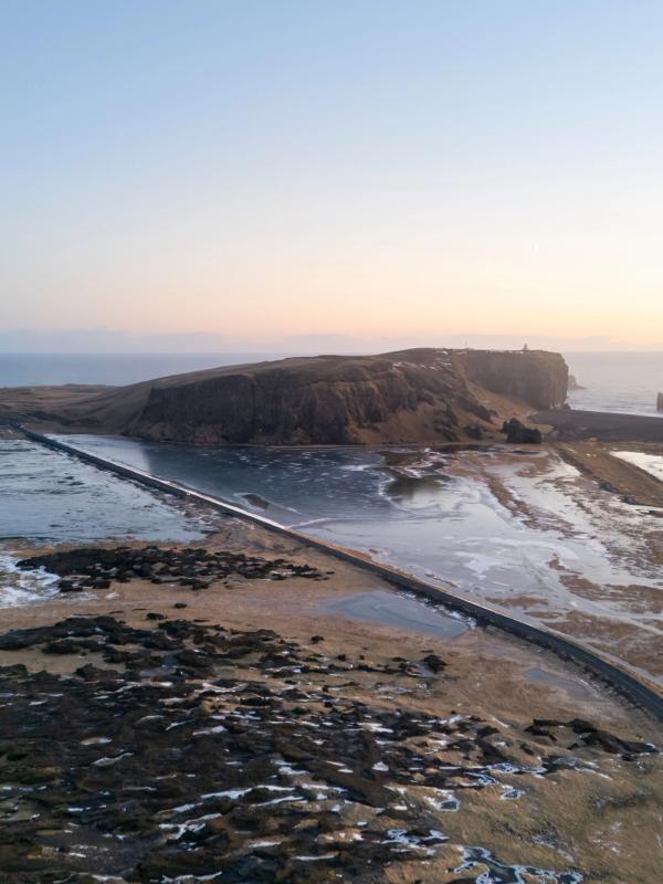 Aerial view of a rugged coastal landscape at dawn or dusk, with a road crossing a partially frozen lagoon towards a dark cliff topped by a lighthouse and sea stacks in the misty distance.