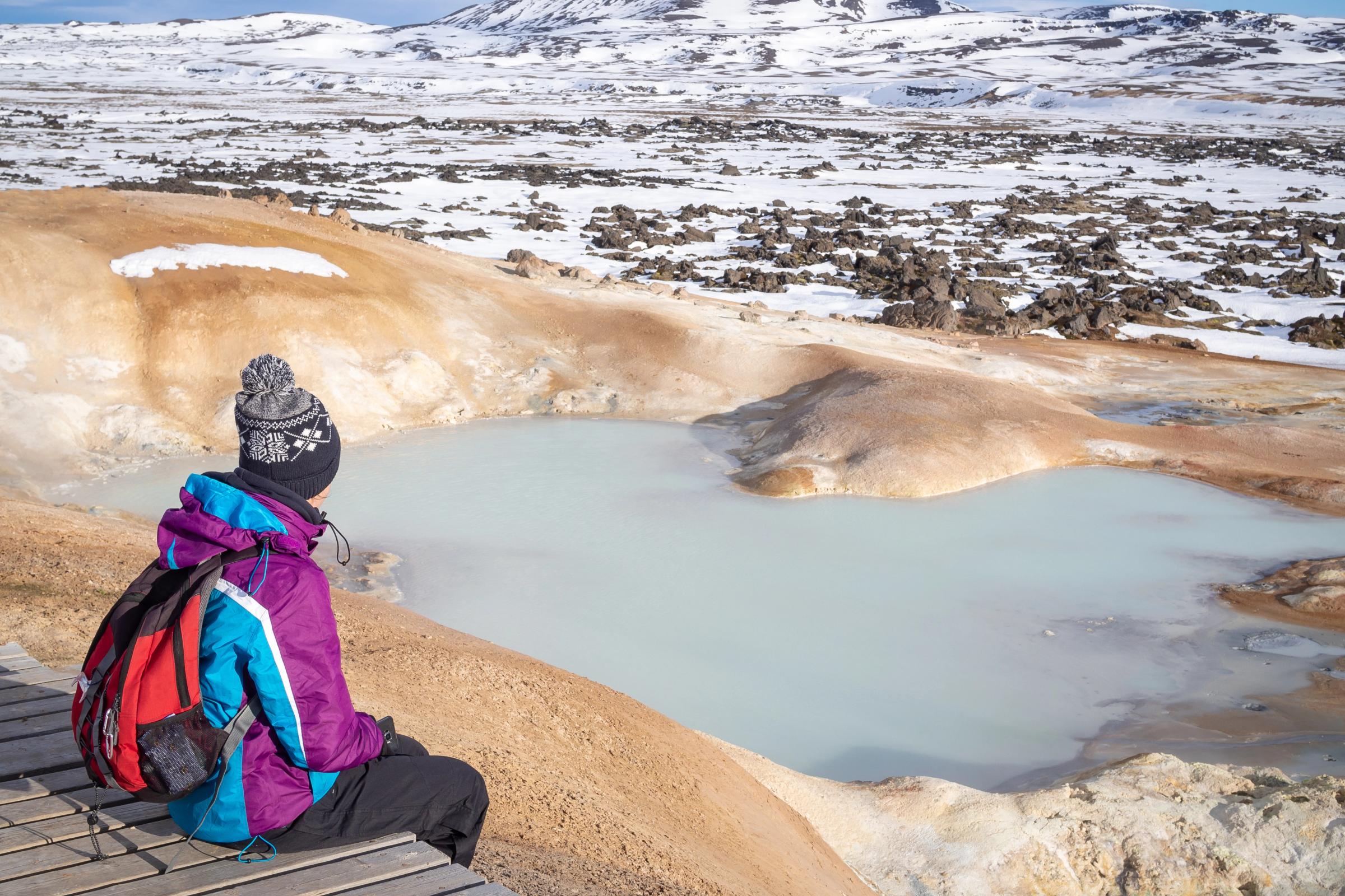 A person in a winter jacket and hat sits on a boardwalk, looking at a light blue geothermal pool in a snowy, rocky landscape with distant mountains.