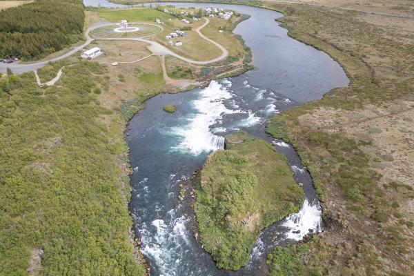 Aerial view of a powerful river with several waterfalls flowing past green land, a campsite, and a circular structure.