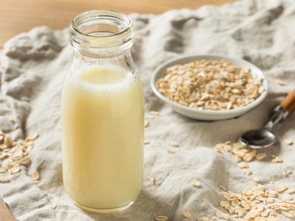 A bottle of oat milk with a bowl of rolled oats and a spoon on a cloth.