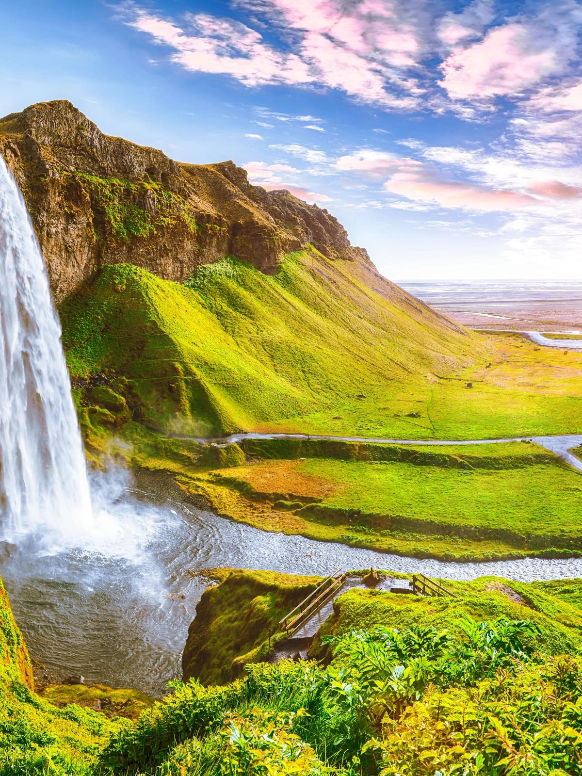 A waterfall cascades down a cliff into a lush green valley with a winding river under a bright sky.